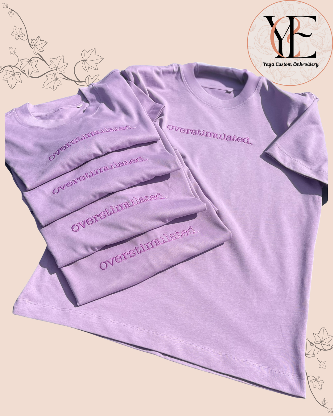 Stack of lavender t-shirts with 'Overstimulated' text embroidered on the front on a beige background