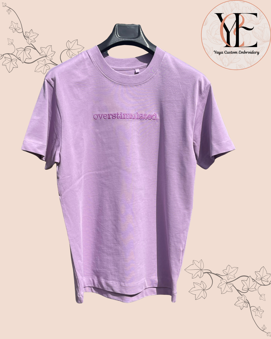 Purple t-shirt with overstimulated text embroidered on the front on a beige background