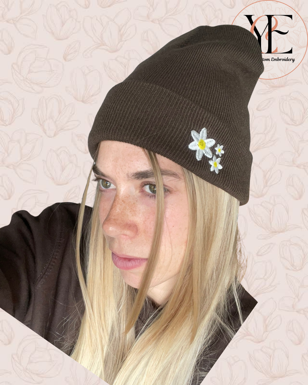 Person wearing a brown beanie with 3 white daisies embroidery on a textured beige background