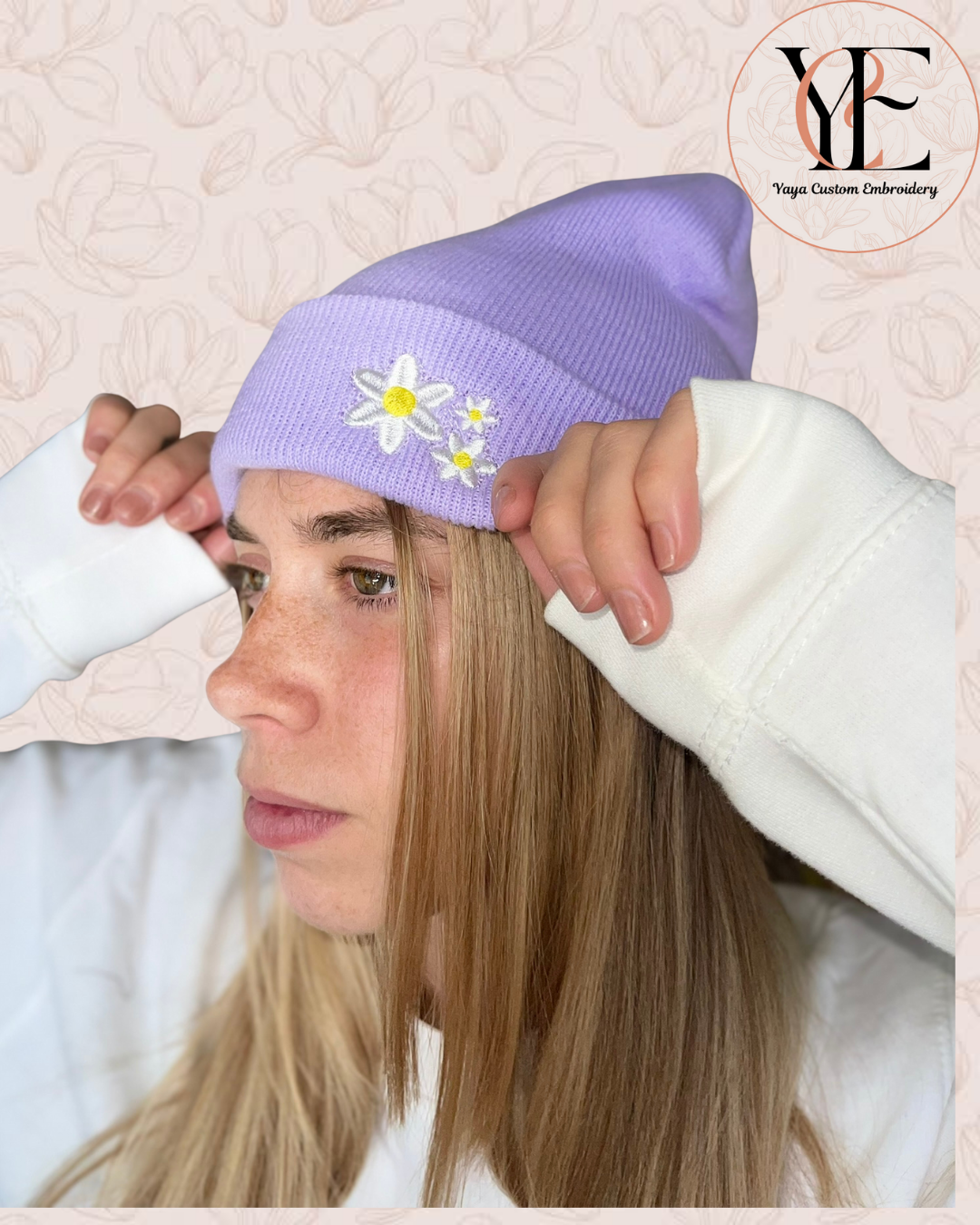 Person wearing a light purple beanie with floral designs, held by hands against a textured beige background.