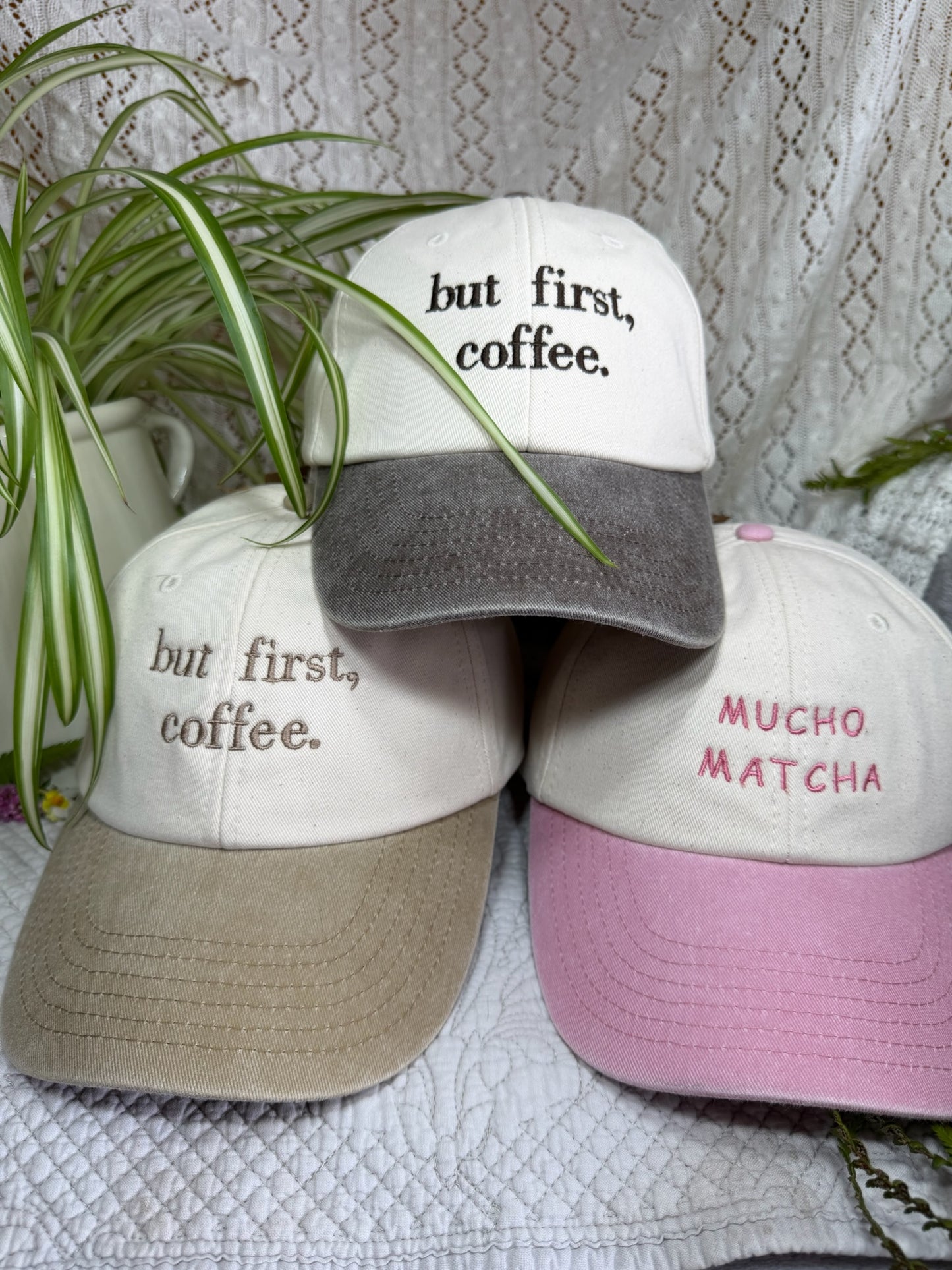 Three baseball caps arranged together on a textured white fabric, with green plant leaves draped over them. The caps have embroidered text: the top cap reads 'but first, coffee.