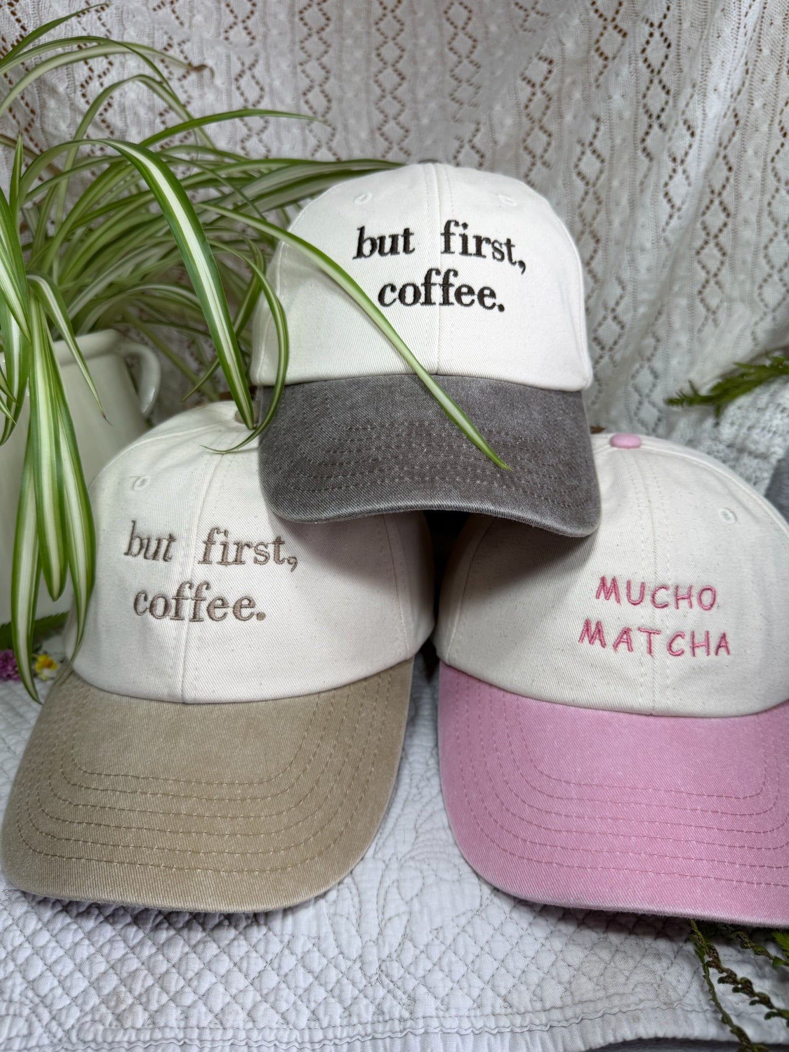 Three baseball caps arranged together on a textured white fabric, with green plant leaves draped over them. The caps have embroidered text: the top cap reads 'but first, coffee.