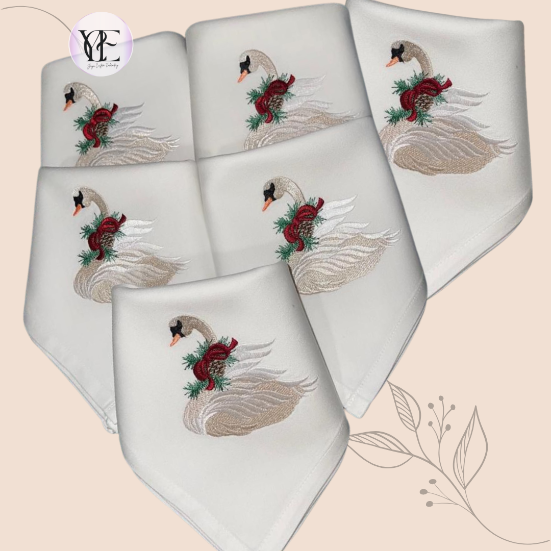 6 folded white napkins showing the embroidered swan with a burgundy ribbon and pine cone around the swans neck.