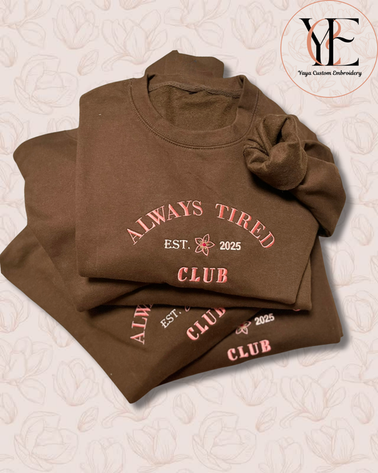Folded brown sweatshirt with 'Always Tired Club' text embroidered using pink letters on a elegant design