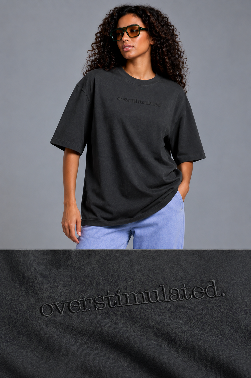 A woman wearing an oversized black t-shirt with tonal embroidered 'overstimulated.' text on the chest, paired with light blue jeans, against a solid gray studio background.