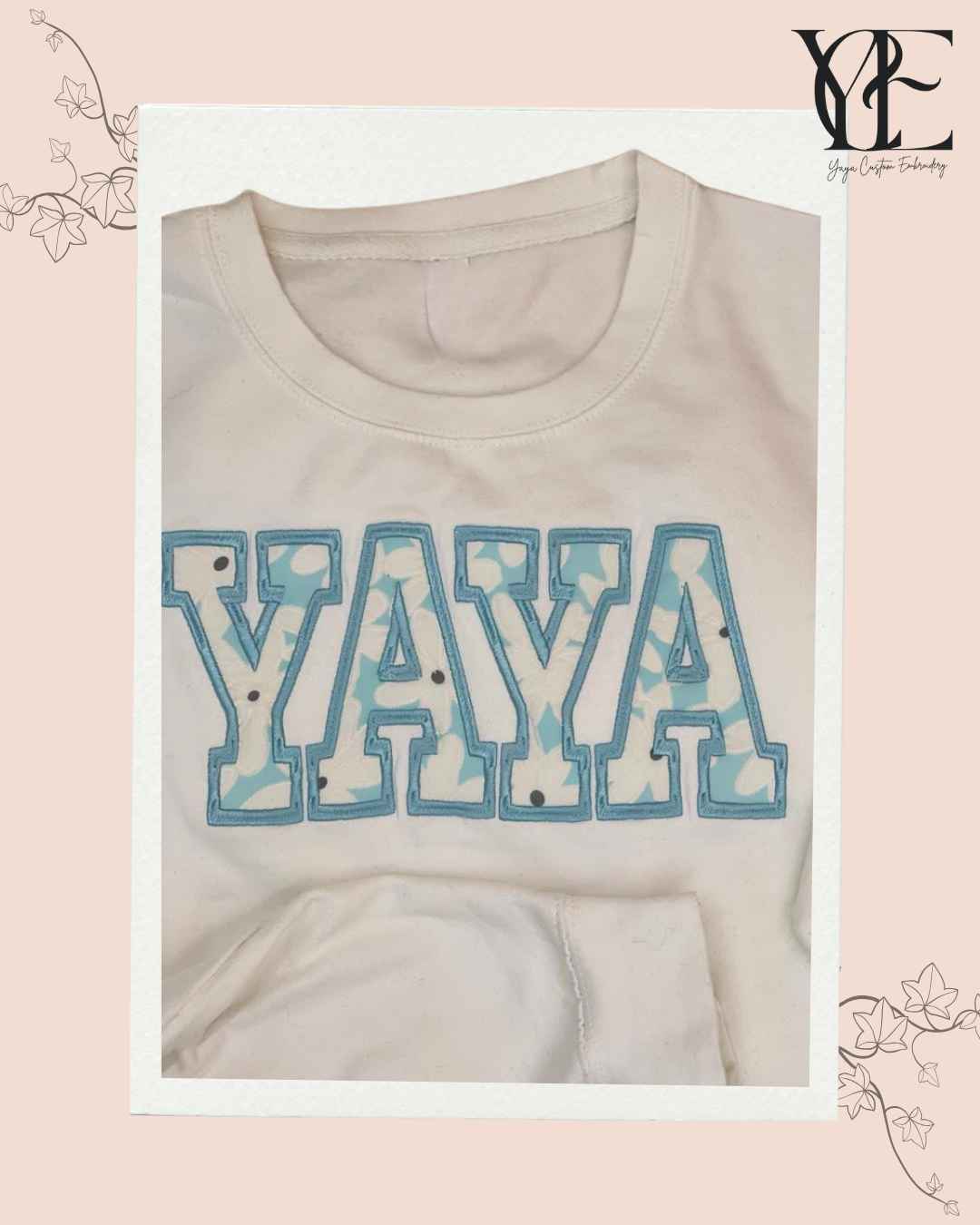 Close up of the YAYA embroidery of a white sweatshirt using college font applique with a baby blue outside satin stitch and a delicate white daisies over a light blue material for the applique. By Yaya Custom Embroidery