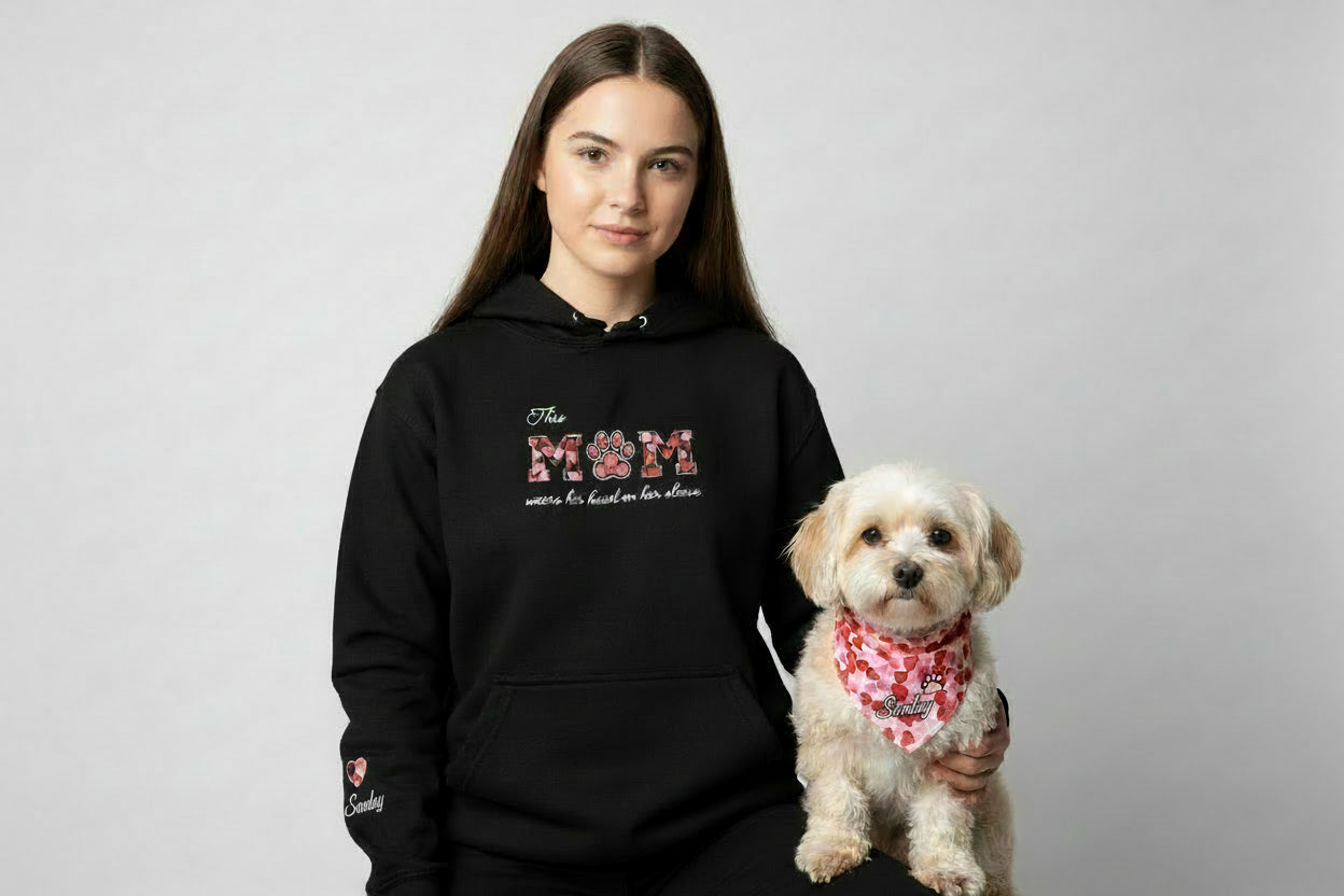 Lady wearing a black hoodie with MUM applique and the slogan "This MUM wears her heart on her sleeve" embroidered on the chest where the letter U in MUM has the shape of a paw. The applique material are hearts on a black background for the M and white for the paw. On the sleeve is the name of the dog, Sunday, and an applique heart too. The dog is wearing a matching heart pattern bandana with her name and a paw embroidered.