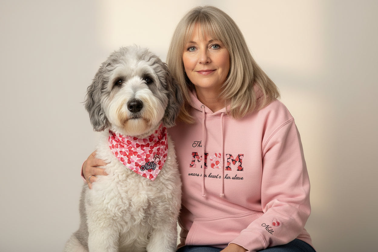 A lady and a dog sitting. The Lady wears a pink hoodie with the embroidery This MUM wears her heart on her sleeve. On her sleeve there's an embroidery of the dog's name and a heart matching the dog's bandana and the applique MUM letters of the chest. The letter U has the shape of a dog's paw.