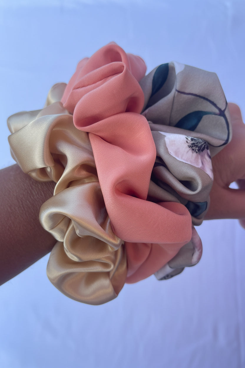 A close-up of a wrist wearing a set of three satin hair scrunchies. The scrunchies are in three distinct colors: a gold satin, a dusty pink satin, and a taupe-beige satin with a dark green leaf pattern and a small white floral accent.