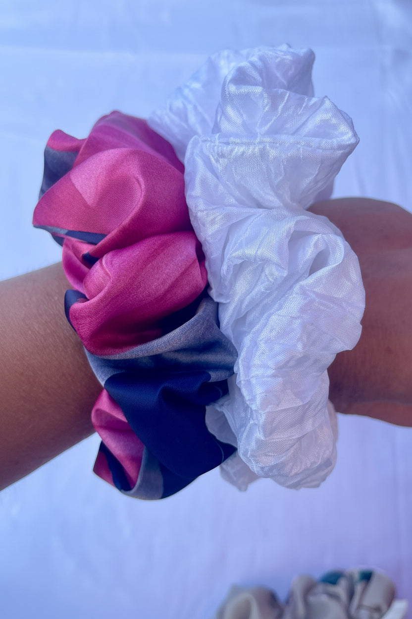 A close-up of a fabric hair scrunchie worn on a wrist, showing two main fabric sections: one in shiny pink and navy blue, and one in sheer white.