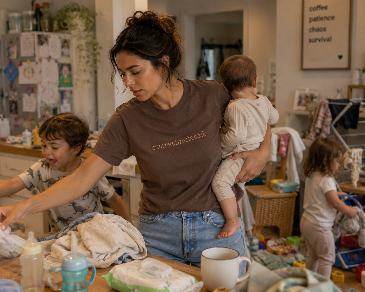A woman in a chocolate brown t-shirt with the text 'overstimulated.' embroidered on the chest holds a baby in a busy home setting with children and household items in the background.