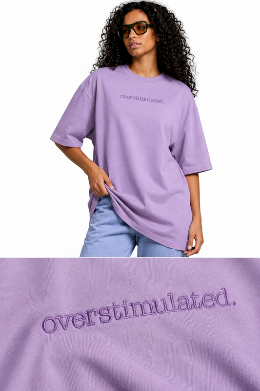 A model wearing an oversized lavender t-shirt with 'overstimulated.' embroidered on the chest, paired with light blue jeans, against a solid gray studio background.
