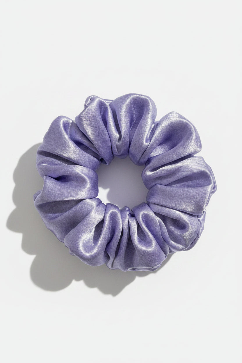 A single satin hair scrunchie in a shimmering lavender-purple color, photographed close-up against a light fabric background.