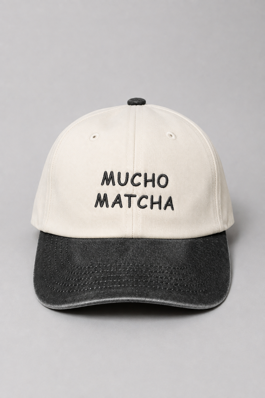 A soft unstructured crown baseball cap with an off-white crown and a black curved brim. The front of the cap features the words 'MUCHO MATCHA' embroidered in black.