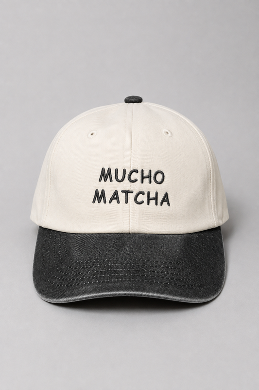 A soft unstructured crown baseball cap with an off-white crown and a black curved brim. The front of the cap features the words 'MUCHO MATCHA' embroidered in black.