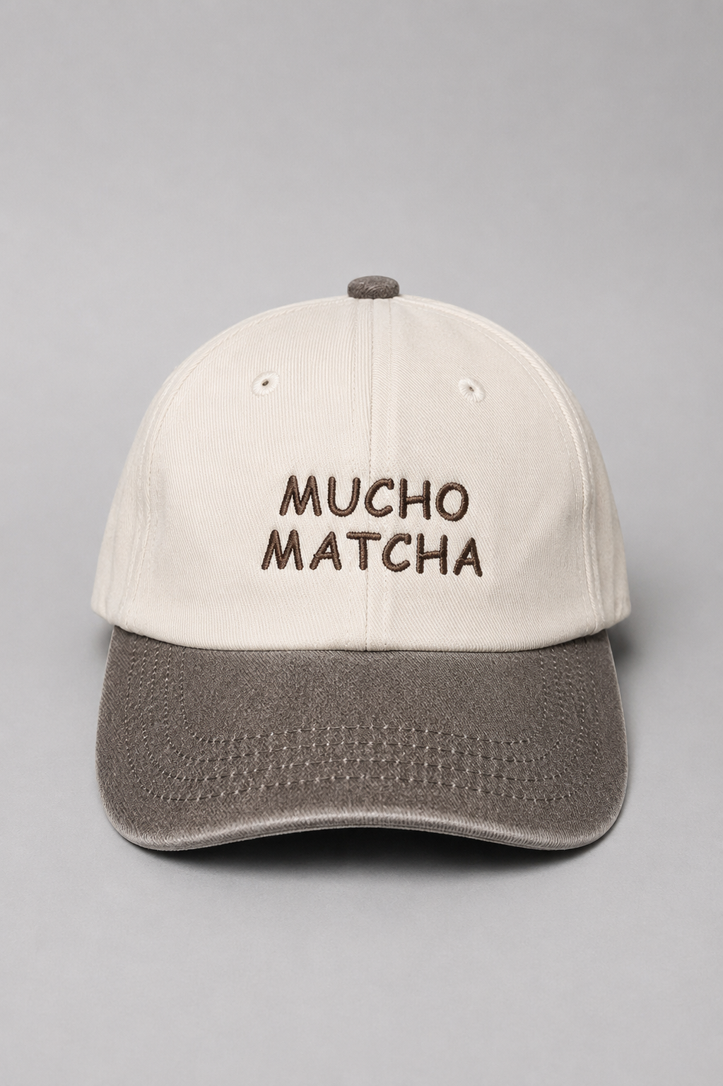 A soft unstructured crown baseball cap with an off-white crown and a brown curved brim. The front of the cap features the words 'MUCHO MATCHA' embroidered in brown.