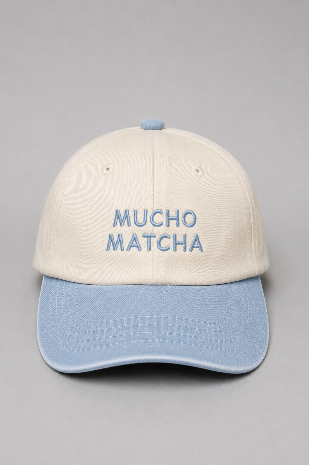 A soft unstructured crown baseball cap with an off-white crown and a light blue curved brim. The front of the cap features the words 'MUCHO MATCHA' embroidered in blue.