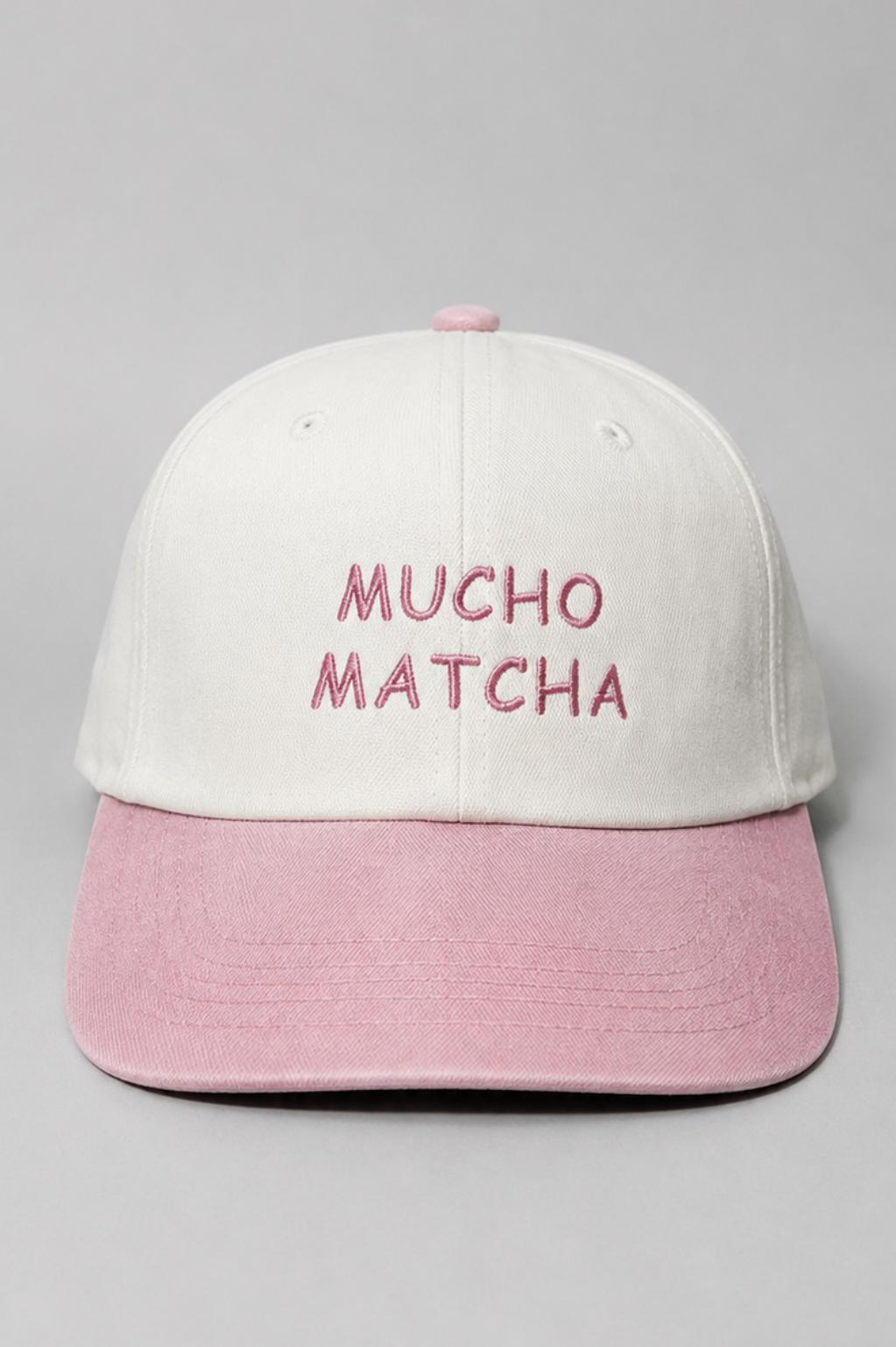 A soft unstructured crown baseball cap with an off-white crown and a light pink curved brim. The front of the cap features the words 'MUCHO MATCHA' embroidered in pink.