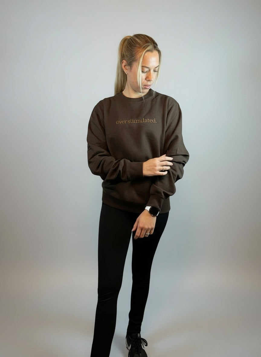 Lady standing wearing a chocolate sweatshirt with the word "Overstimulated." embroidered over the chest on a lighter chocolate thread 