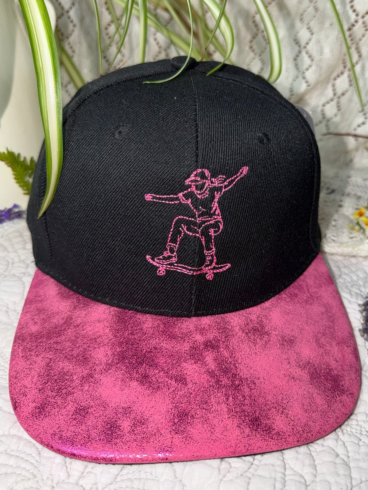 A black flat-brim snapback cap with a bright pink marbled brim and a pink embroidered outline of a person skateboarding on the front, styled with green plants on a white textured fabric background.