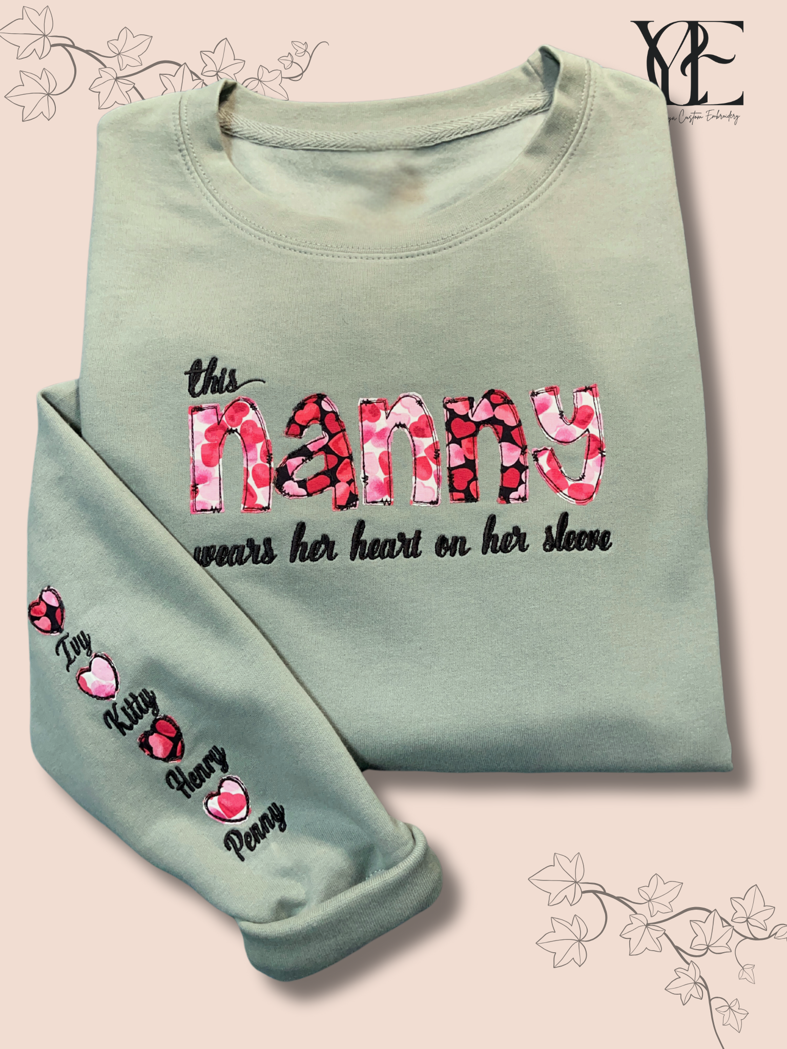 Folded dusty green sweatshirt embroidered  on the chest with "This nanny wears her heart on her sleeve" while the sleeve has the names of her grandchildren.