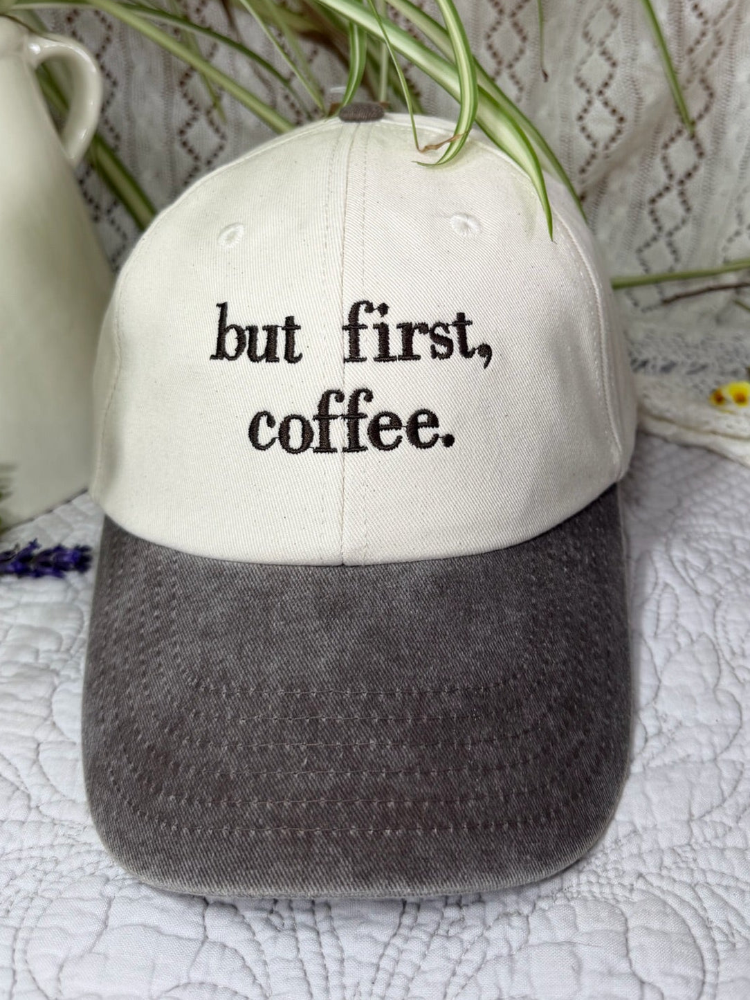 A baseball cap with a cream-colored crown and a dark gray textured visor, featuring the text 'but first, coffee.' embroidered on the front.