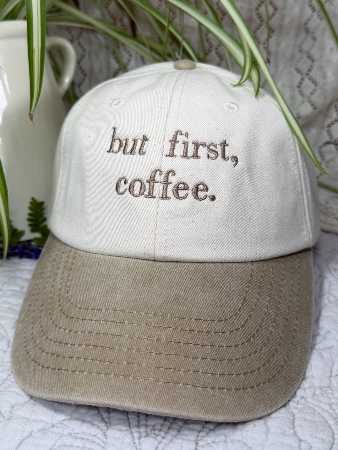 A baseball cap with a cream-colored crown and a taupe-beige curved brim, featuring the embroidered text 'but first, coffee.' in brown.