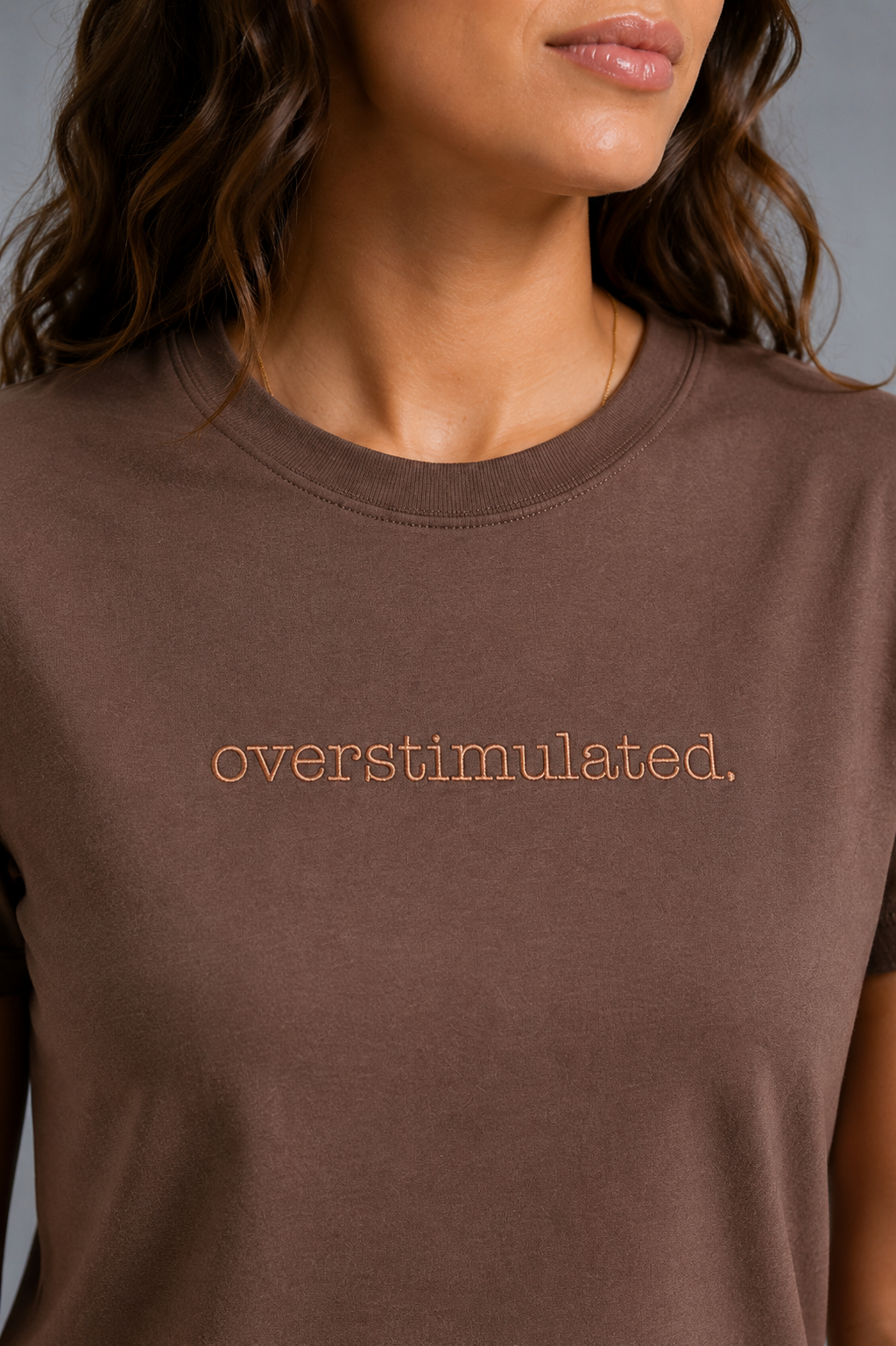 A close-up of a woman wearing a chocolate brown crewneck t-shirt with the word 'overstimulated.' embroidered  in a lighter brown lowercase font across the chest.