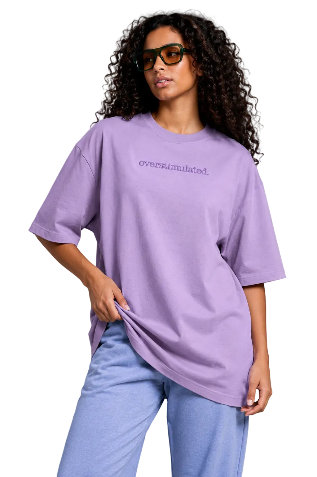 A woman with curly hair wearing an oversized lavender t-shirt with the word 'overstimulated.' embroidered in purple on the chest, paired with light blue sweatpants, against a solid gray studio background.
