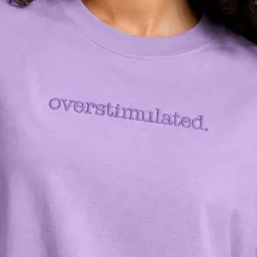 A close-up of a person wearing a lavender purple crewneck sweatshirt with the word 'overstimulated.' embroidered in matching purple across the chest.