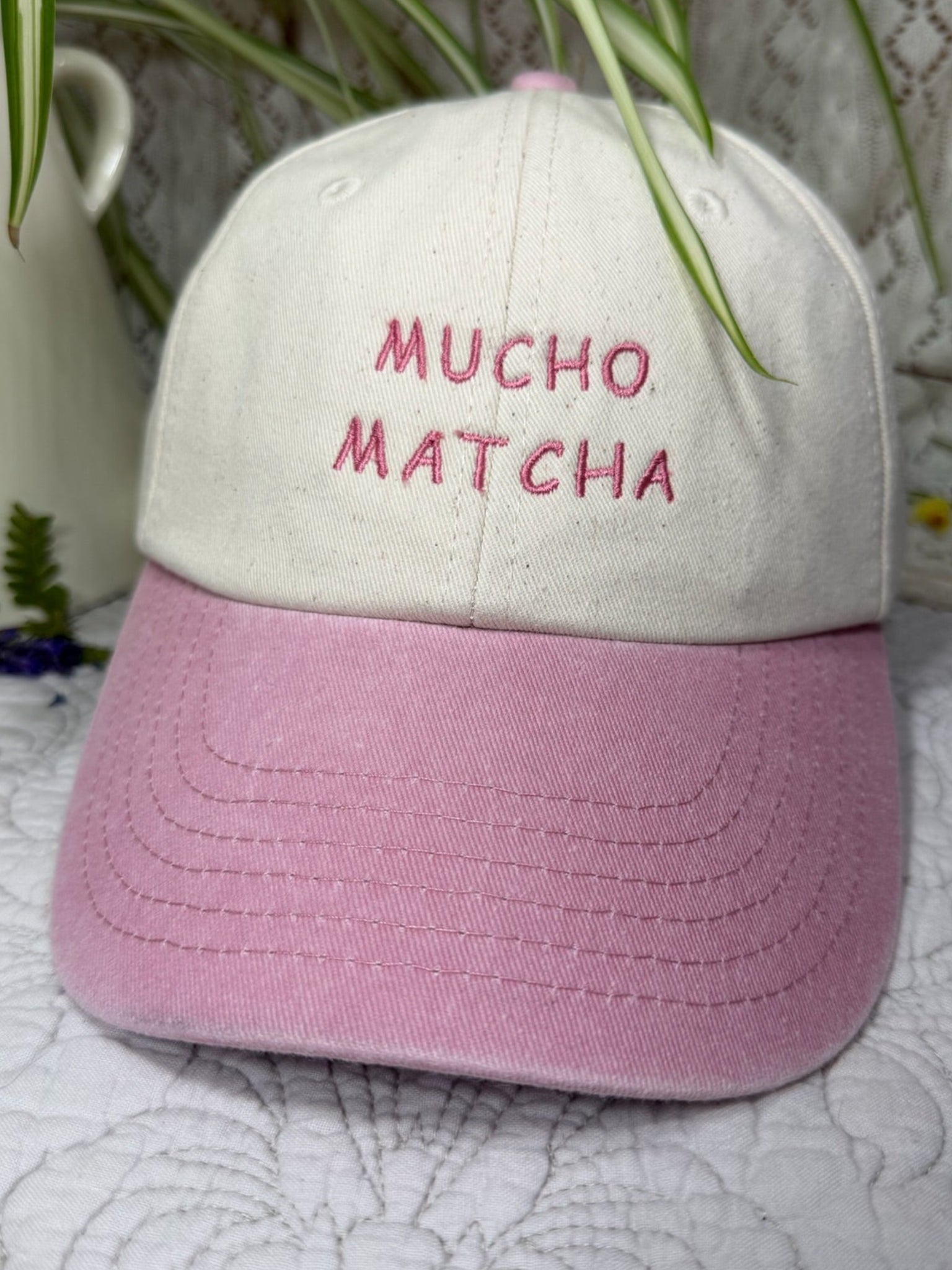 A baseball cap with an off-white crown and a pink brim, featuring the words 'MUCHO MATCHA' embroidered in pink on the front. The cap is styled on a white patterned fabric surface with a white ceramic pitcher and green plant leaves in the background.
