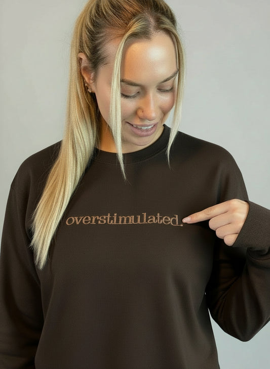 Lady wearing a chocolate brown overstimulated sweatshirt and she is pointing to the embroidered word
