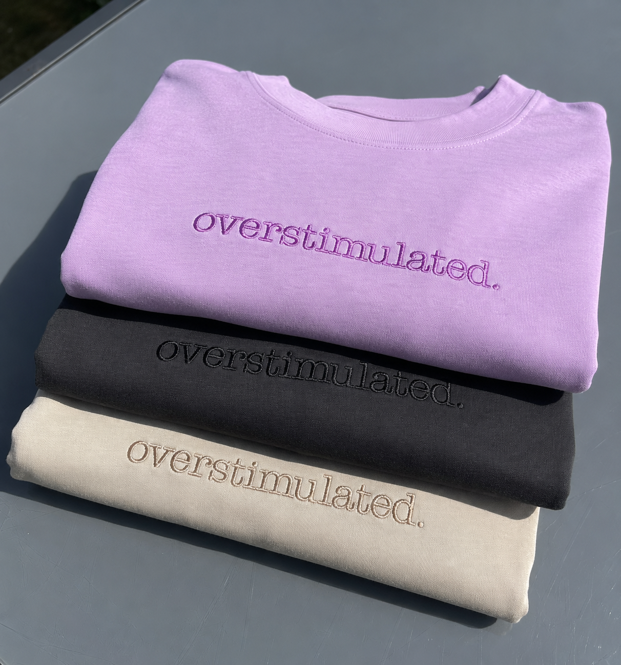 Three folded crewneck T-shirt stacked on a gray surface, each in a different colour with the word 'overstimulated.' embroidered across the chest in matching thread.